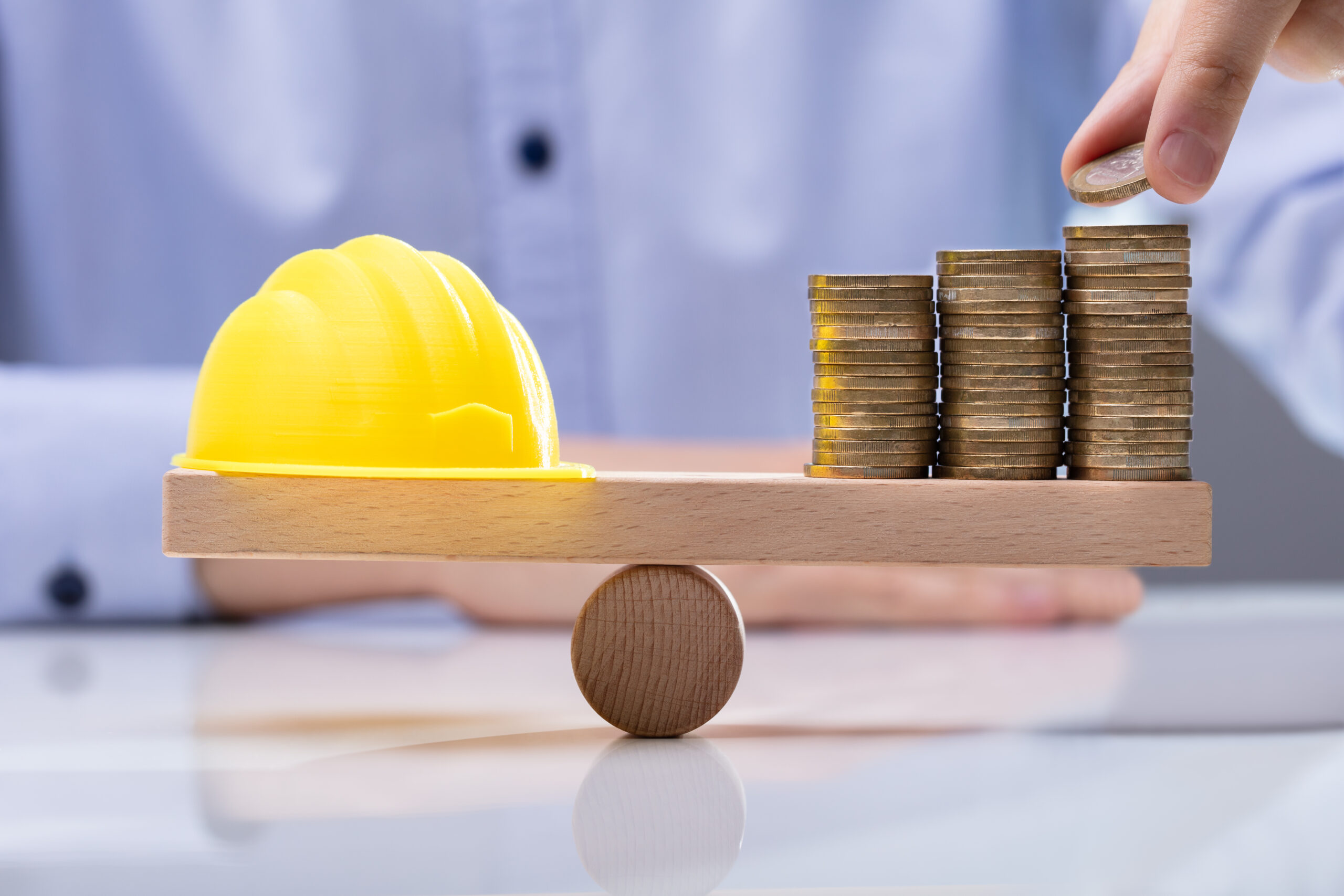 Yellow Hard Hat And Stack Of Coins On Seesaw WORKERS COMPENSATION