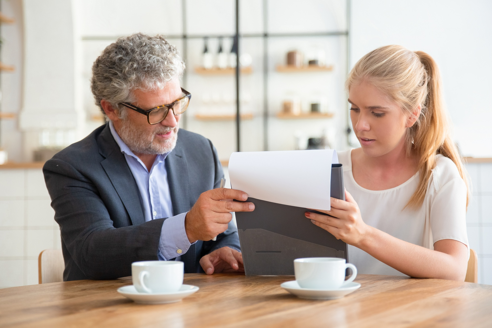 An older man in a suit and glasses sits at a table with a young woman, pointing at documents she is holding. Both are focused on the papers—likely discussing wills and estate matters. Two coffee cups are on the table in front of them. An older man in a suit and glasses sits at a table with a young woman, pointing at documents she is holding. Both are focused on the papers—likely discussing wills and estate matters. Two coffee cups are on the table in front of them.