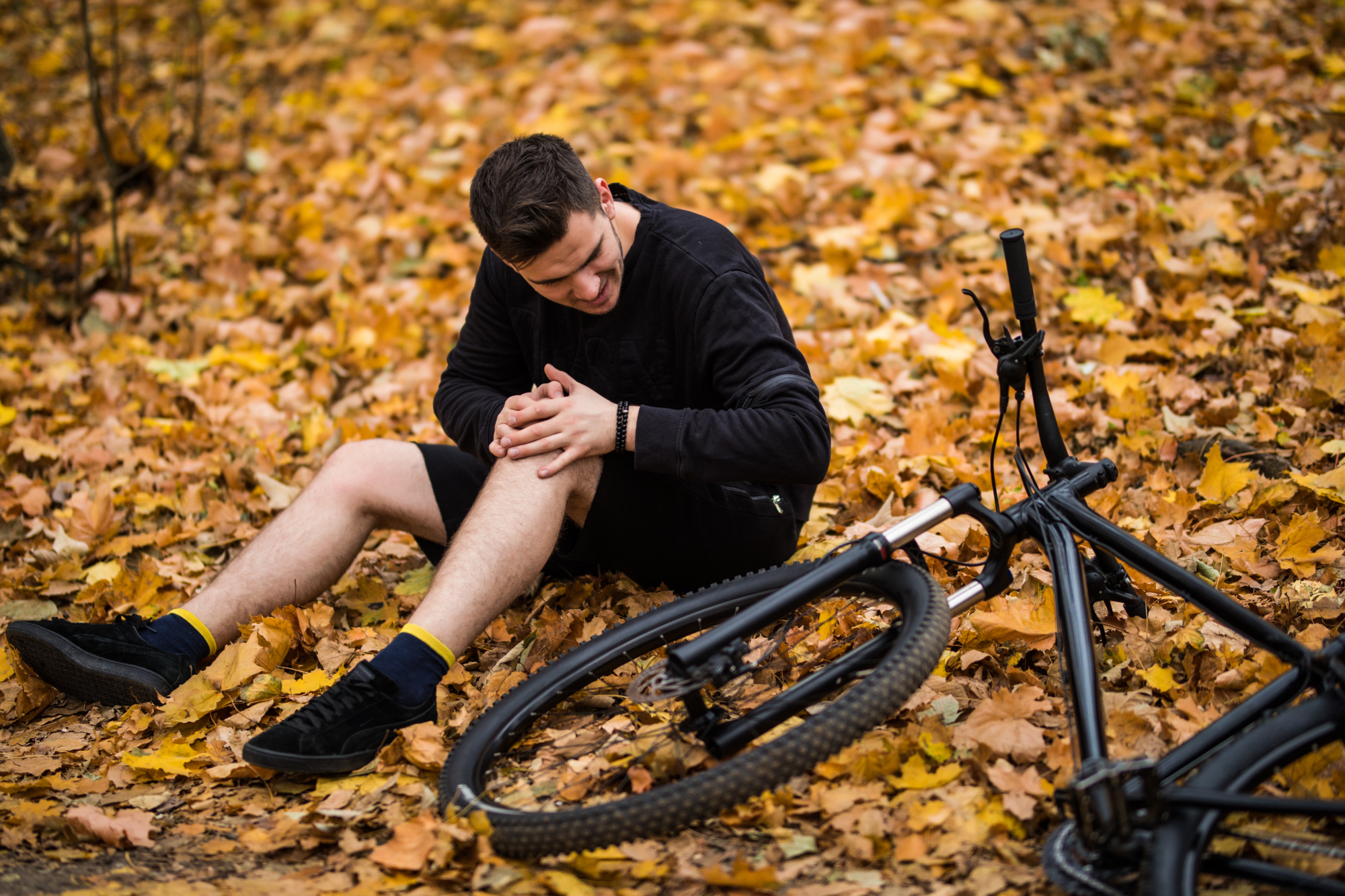 A man sits on the ground covered with autumn leaves, holding his knee in pain next to a fallen bicycle—a typical scene of cyclist accidents. A man sits on the ground covered with autumn leaves, holding his knee in pain next to a fallen bicycle—a typical scene of cyclist accidents.