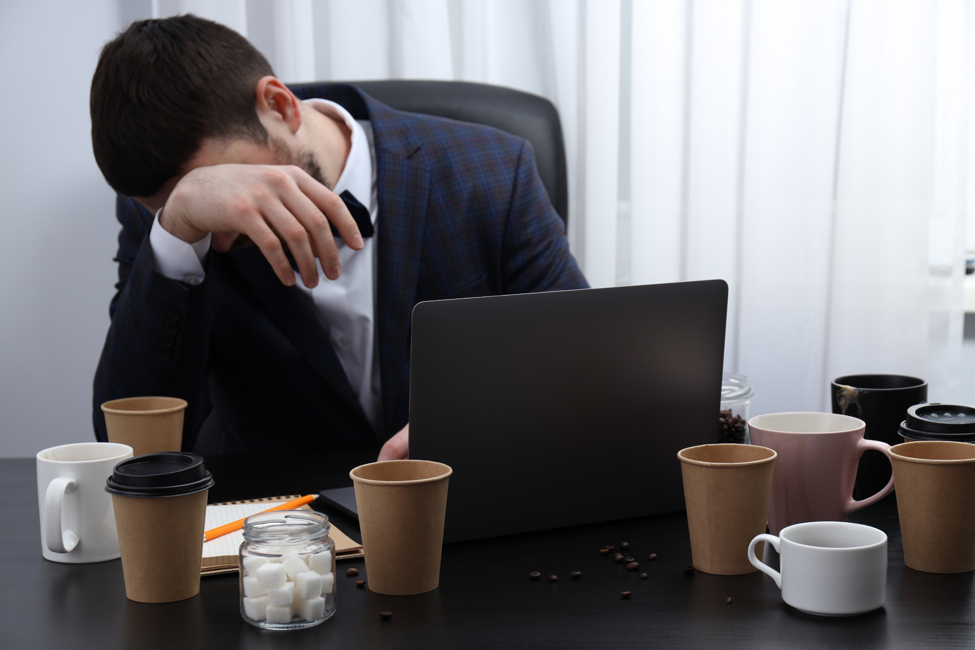A man in a suit sits at a desk, resting his head on his hand in frustration as he faces superannuation paperwork. His laptop is open, and the desk is cluttered with empty coffee cups, a notebook, and a jar of sugar cubes. A man in a suit sits at a desk, resting his head on his hand in frustration as he faces superannuation paperwork. His laptop is open, and the desk is cluttered with empty coffee cups, a notebook, and a jar of sugar cubes.