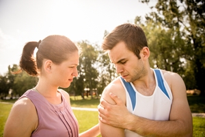 A woman comforts a man outdoors as he holds his shoulder in pain, possibly considering injury compensation. They appear to be in a park with trees and sunlight in the background. A woman comforts a man outdoors as he holds his shoulder in pain, possibly considering injury compensation. They appear to be in a park with trees and sunlight in the background.