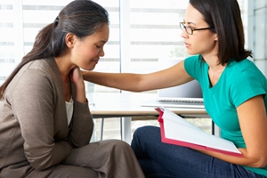 A woman in a brown jacket looks upset while sitting next to another woman in glasses and a teal shirt, who is comforting her about injury compensation with a hand on her shoulder and holding a folder. A woman in a brown jacket looks upset while sitting next to another woman in glasses and a teal shirt, who is comforting her about injury compensation with a hand on her shoulder and holding a folder.