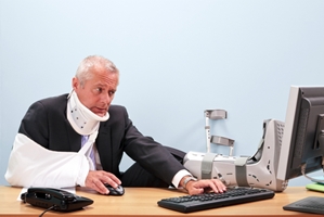 A man in a suit wearing a neck brace, arm sling, and foot cast sits at a desk using a computer, looking frustrated as he deals with workers compensation paperwork. A man in a suit wearing a neck brace, arm sling, and foot cast sits at a desk using a computer, looking frustrated as he deals with workers compensation paperwork.