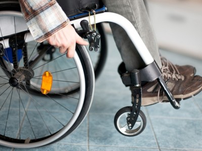 A close-up of a person with total and permanent disability in a wheelchair, wearing brown shoes and gray pants, with one hand on the wheel and the other adjusting the wheel lock on a tiled floor. A close-up of a person with total and permanent disability in a wheelchair, wearing brown shoes and gray pants, with one hand on the wheel and the other adjusting the wheel lock on a tiled floor.