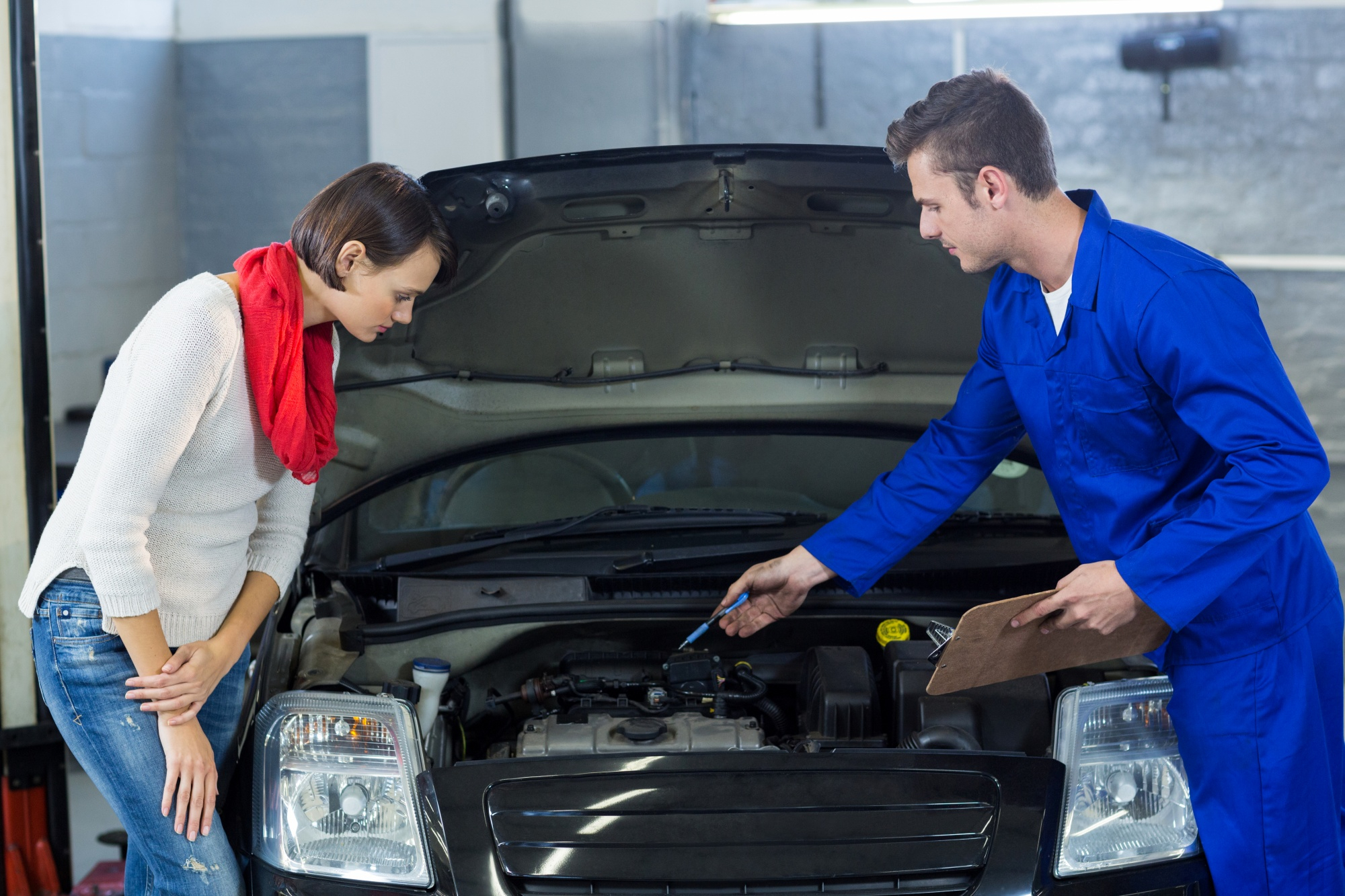 A mechanic in blue coveralls explains something under the open hood of a car to a woman in a white sweater and red scarf, possibly after a car accident Sydney locals might face. The woman leans forward attentively in the garage setting. A mechanic in blue coveralls explains something under the open hood of a car to a woman in a white sweater and red scarf, possibly after a car accident Sydney locals might face. The woman leans forward attentively in the garage setting.