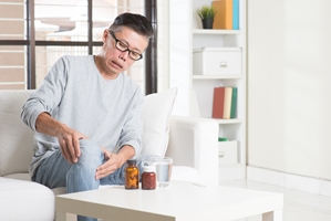 An older man sits on a couch, touching his knee in discomfort. Pill bottles and a glass of water on the table suggest he may be considering medication for pain relief after suffering private property injuries. An older man sits on a couch, touching his knee in discomfort. Pill bottles and a glass of water on the table suggest he may be considering medication for pain relief after suffering private property injuries.