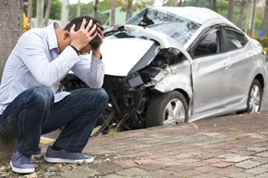 A man sits on the curb with his head in his hands, appearing distressed, in front of a heavily damaged silver car after a car accident. A man sits on the curb with his head in his hands, appearing distressed, in front of a heavily damaged silver car after a car accident.