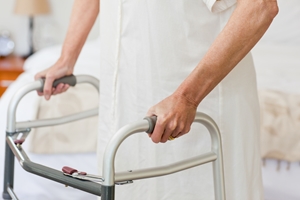 An injured worker wearing a white robe is standing and holding onto a walker for support in a bedroom. Only their hands and part of their body are visible. An injured worker wearing a white robe is standing and holding onto a walker for support in a bedroom. Only their hands and part of their body are visible.