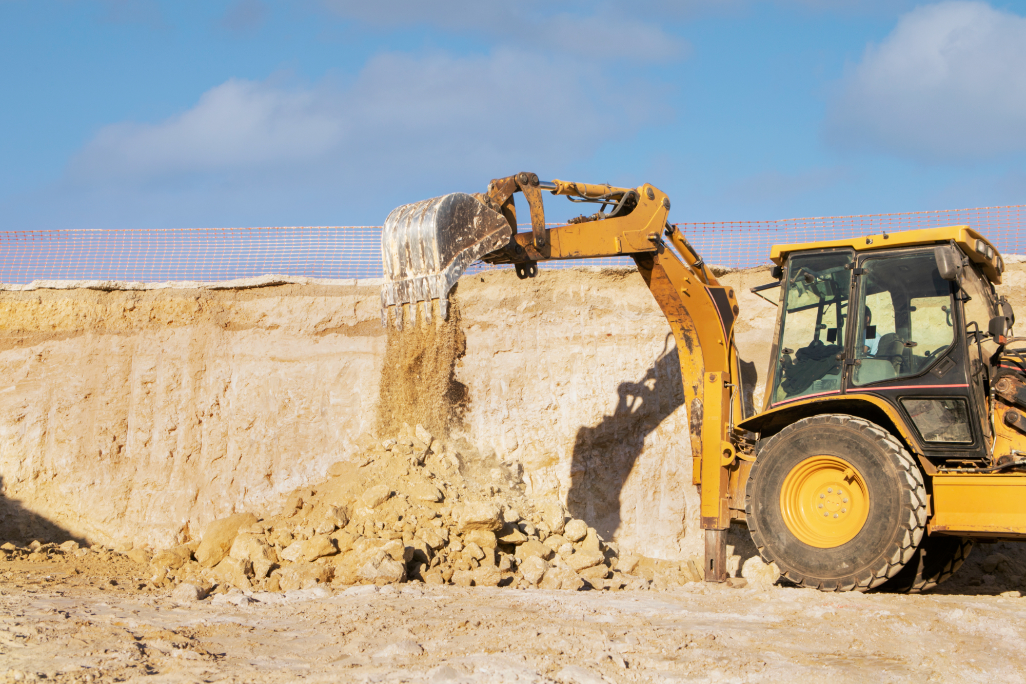 A yellow excavator dumps sand and rocks at a construction site, with a clear sky in the background and a safety fence along the edge of the excavation area, where workers are assured proper compensation for their efforts. A yellow excavator dumps sand and rocks at a construction site, with a clear sky in the background and a safety fence along the edge of the excavation area, where workers are assured proper compensation for their efforts.
