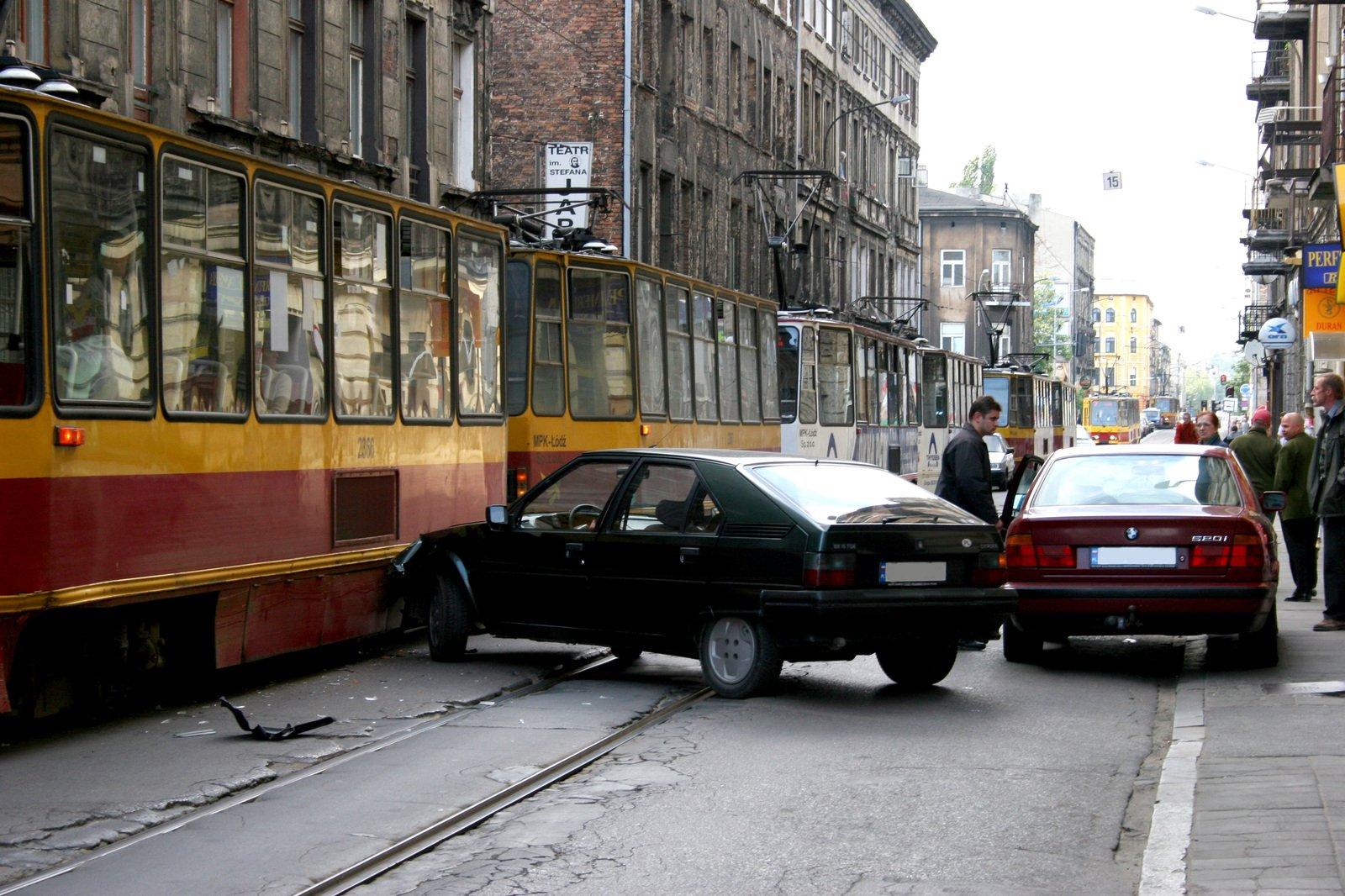 Two cars have collided on a city street, with one car crashed into the side of a yellow tram. People stand nearby observing the scene, and the street is lined with old buildings. Two cars have collided on a city street, with one car crashed into the side of a yellow tram. People stand nearby observing the scene, and the street is lined with old buildings.
