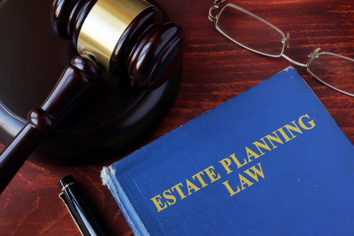 A blue book titled “Estate Planning Law” is on a wooden desk next to a gavel, eyeglasses, and a black pen. A blue book titled "Estate Planning Law" is on a wooden desk next to a gavel, eyeglasses, and a black pen.