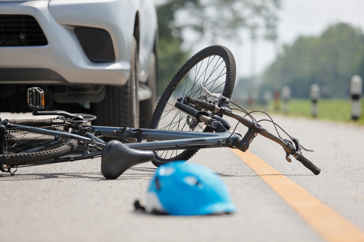 A bicycle lies on its side in the road near a white car, with a blue helmet on the ground, suggesting a recent accident. The scene is on an empty, rural road with trees in the background. Accident car crash with bicycle on road