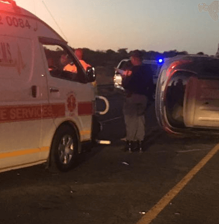 An ambulance and emergency personnel attend the scene of a car accident at dusk, with one vehicle overturned on its side on the road. Blue and red emergency lights are visible in the background. An ambulance and emergency personnel attend the scene of a car accident at dusk, with one vehicle overturned on its side on the road. Blue and red emergency lights are visible in the background.
