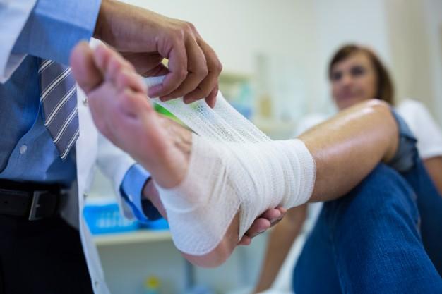 A medical professional wraps a bandage around a patient’s injured foot while the patient sits on an exam table in a clinic. A medical professional wraps a bandage around a patient's injured foot while the patient sits on an exam table in a clinic.