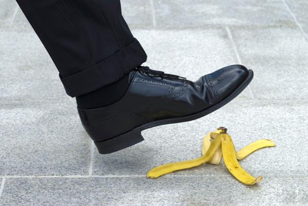 A person wearing black dress shoes and dark pants is about to step on a yellow banana peel on a gray tiled floor. A person wearing black dress shoes and dark pants is about to step on a yellow banana peel on a gray tiled floor.
