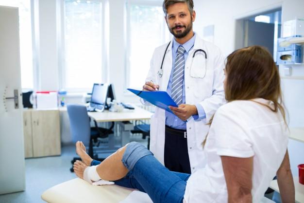 A doctor with a stethoscope and clipboard talks to a patient with a bandaged knee, who is sitting on an examination table in a medical office. A doctor with a stethoscope and clipboard talks to a patient with a bandaged knee, who is sitting on an examination table in a medical office.