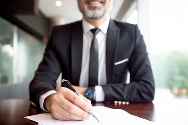 A man in a suit sitting at a desk, smiling, and holding a pen while writing on a sheet of paper. A man in a suit sitting at a desk, smiling, and holding a pen while writing on a sheet of paper.