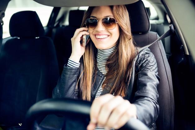 A smiling woman wearing sunglasses and a leather jacket is talking on her phone while driving a car. She has one hand on the steering wheel and appears cheerful. A smiling woman wearing sunglasses and a leather jacket is talking on her phone while driving a car. She has one hand on the steering wheel and appears cheerful.
