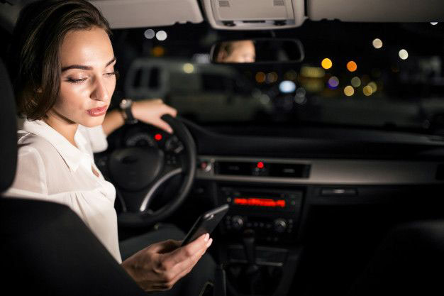 A woman sitting in a car at night looks at her phone while her hand rests on the steering wheel. The car interior is dimly lit, and city lights are visible through the windshield. A woman sitting in a car at night looks at her phone while her hand rests on the steering wheel. The car interior is dimly lit, and city lights are visible through the windshield.