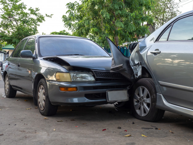Two cars involved in a rear-end collision; the front of a dark sedan is damaged after crashing into the back of a silver car, which also shows damage. Trees and pavement are visible in the background.