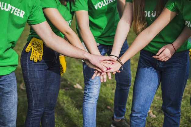Five people wearing green "Volunteer" shirts stand in a circle outdoors and stack their hands together in the center, symbolizing teamwork and unity. The grass is visible underfoot.
