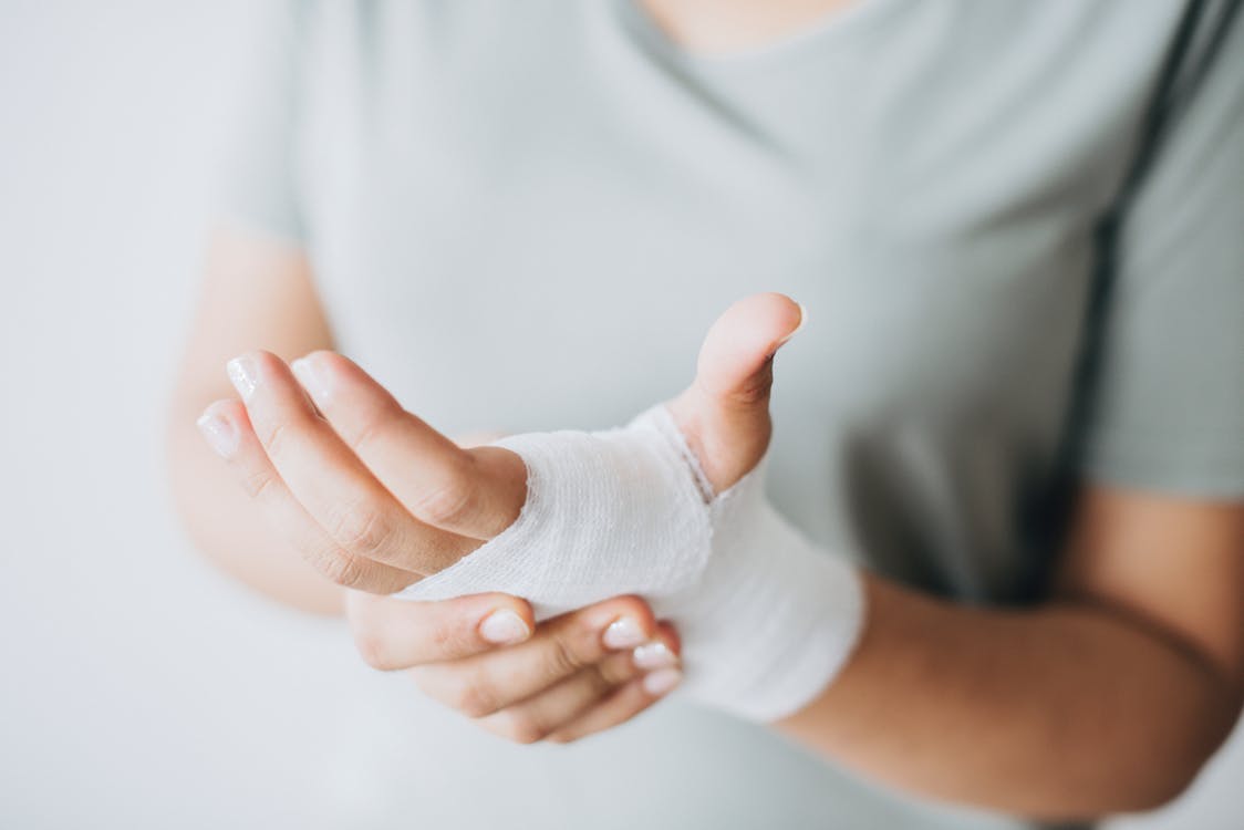 A person wearing a light grey shirt is shown with a bandaged hand and wrist, gently holding their injured hand with the other hand. The background is softly blurred. A person wearing a light grey shirt is shown with a bandaged hand and wrist, gently holding their injured hand with the other hand. The background is softly blurred.