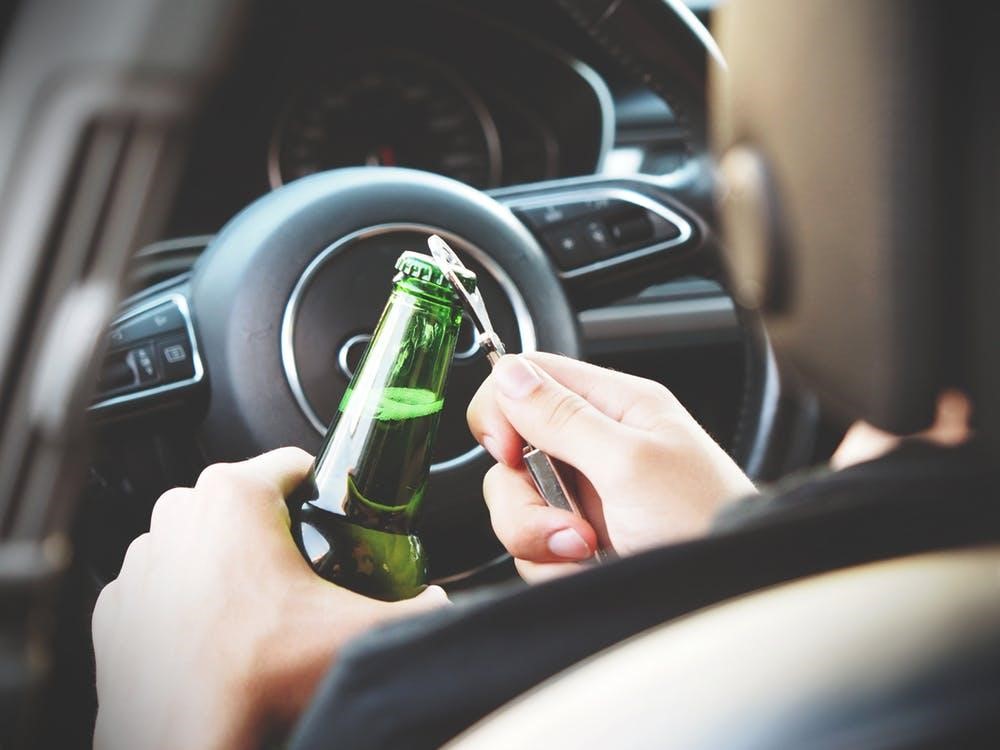 A person sitting in a car uses a bottle opener to open a green beer bottle, holding it near the steering wheel with the dashboard visible in the background. A person sitting in a car uses a bottle opener to open a green beer bottle, holding it near the steering wheel with the dashboard visible in the background.