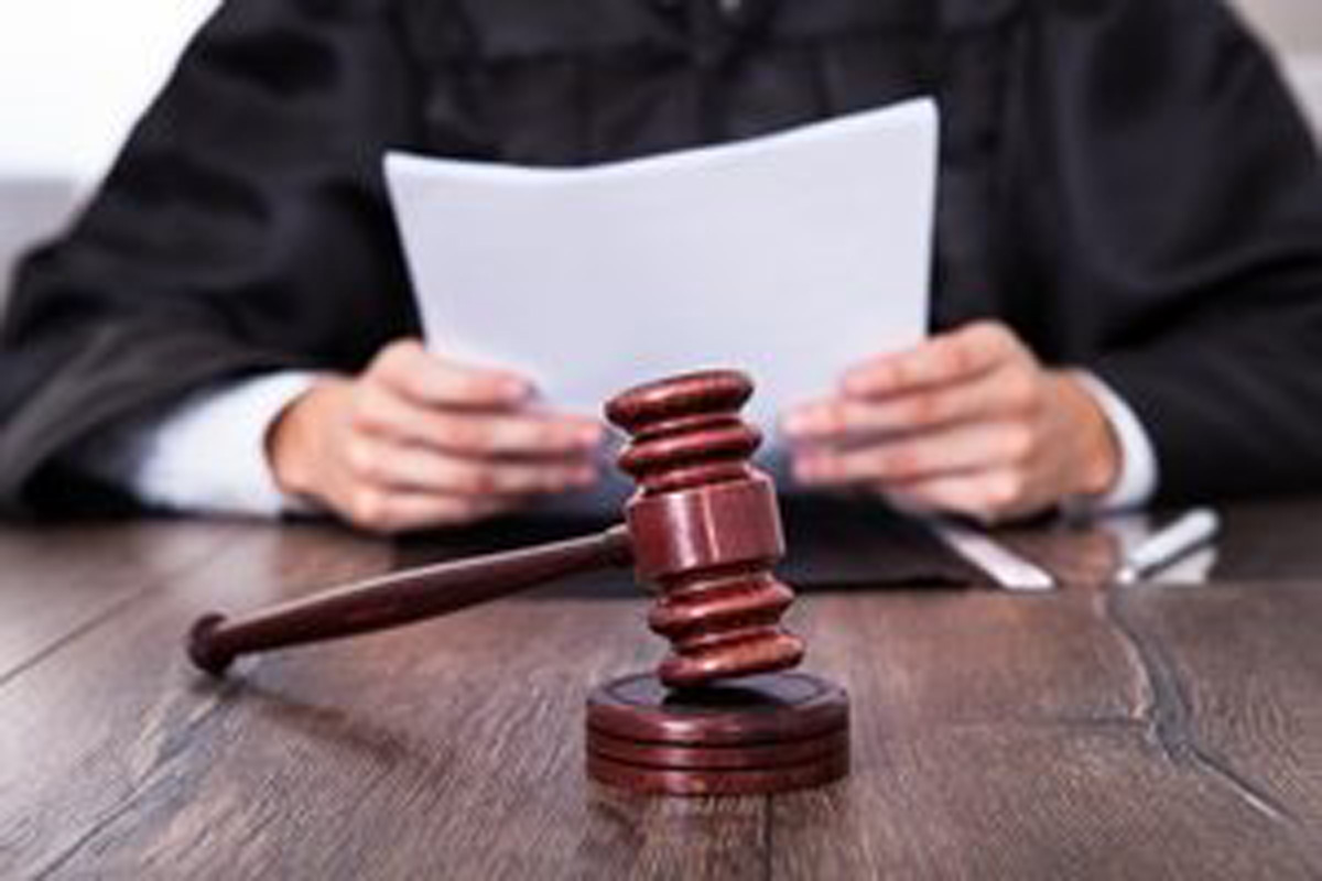 A judge in a black robe holds documents while sitting at a desk, with a wooden gavel and sound block prominently placed in the foreground. A judge in a black robe holds documents while sitting at a desk, with a wooden gavel and sound block prominently placed in the foreground.