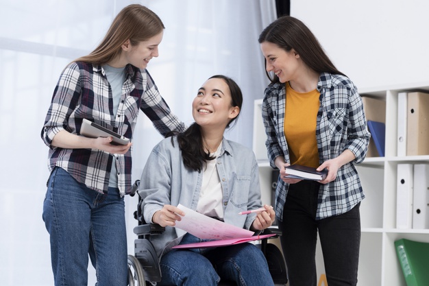 Three young women, one in a wheelchair holding papers, smile and talk together indoors. Two others stand beside her, holding books and a tablet, showing a supportive and friendly atmosphere. Three young women, one in a wheelchair holding papers, smile and talk together indoors. Two others stand beside her, holding books and a tablet, showing a supportive and friendly atmosphere.