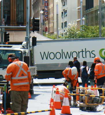 Construction workers in orange safety gear work on a city street near orange cones and machinery, with a Woolworths truck and several pedestrians visible in the background. Construction workers in orange safety gear work on a city street near orange cones and machinery, with a Woolworths truck and several pedestrians visible in the background.