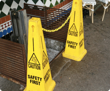 Two yellow “Caution: Safety First” signs and a yellow chain block access to an open stairwell or hole on a sidewalk near a tiled wall, warning pedestrians of a potential hazard. Two yellow "Caution: Safety First" signs and a yellow chain block access to an open stairwell or hole on a sidewalk near a tiled wall, warning pedestrians of a potential hazard.