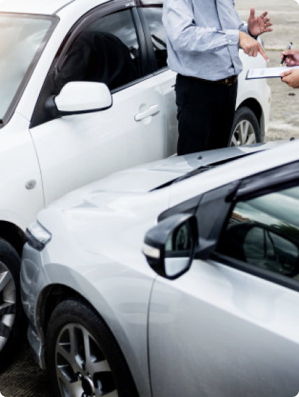 Two people stand between two cars, examining paperwork, likely discussing a car accident or insurance claim. The image is focused on the cars and the individuals' hands holding documents and a pen.