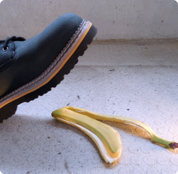 A black shoe is about to step on a yellow banana peel lying on a textured, light-colored floor.