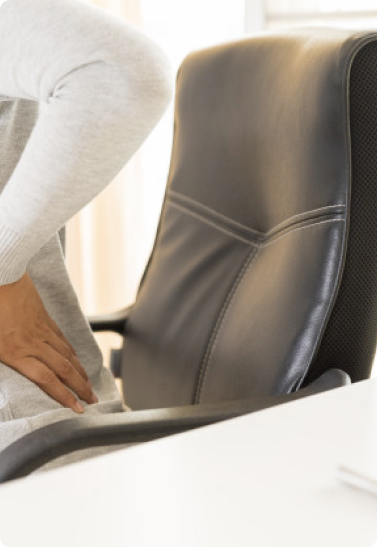 A person wearing a light sweater holds their lower back in discomfort while standing next to a black office chair and a white desk, suggesting back pain possibly from sitting.