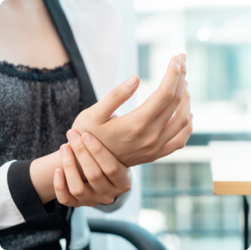 A person in business attire sits indoors, holding their left wrist with their right hand, appearing to experience wrist pain or discomfort.
