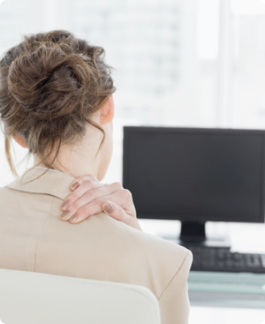 A woman with brown hair in a bun sits at a desk in front of a computer, seen from behind, with one hand on her shoulder, suggesting discomfort or pain.