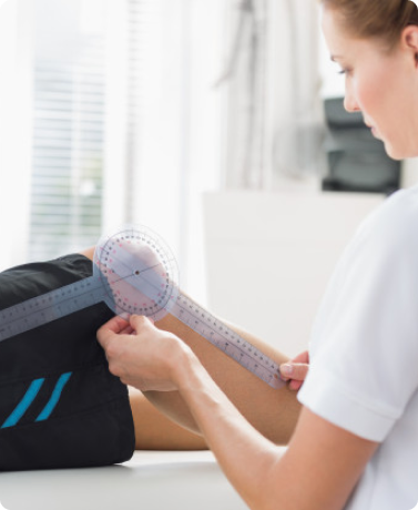 A healthcare professional uses a goniometer to measure the knee joint angle of a patient wearing black shorts with blue stripes, assessing range of motion in a clinical setting.