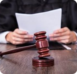 A judge in a black robe holds papers at a desk with a wooden gavel and sound block in the foreground. A judge in a black robe holds papers at a desk with a wooden gavel and sound block in the foreground.