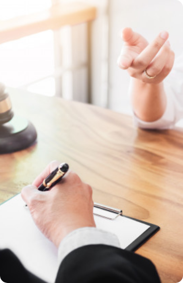 Two people sit at a wooden table; one is writing on a clipboard, while the other gestures with their hand. A gavel is visible on the table, suggesting a legal or professional consultation. Two people sit at a wooden table; one is writing on a clipboard, while the other gestures with their hand. A gavel is visible on the table, suggesting a legal or professional consultation.