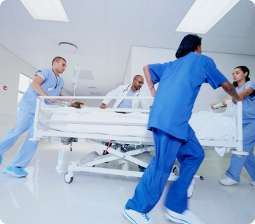 Four medical professionals in blue scrubs and a white coat rush a patient on a hospital bed down a brightly lit hallway, indicating an emergency situation.