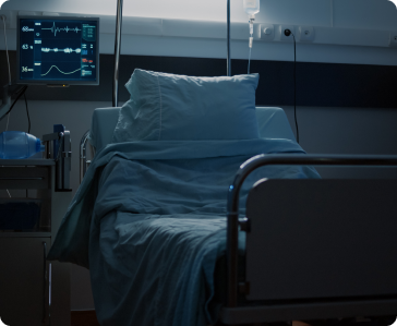 A hospital bed with blue sheets sits empty in a dimly lit room, next to medical equipment and a monitor displaying vital signs.