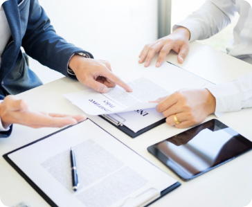 Two people in business attire sit at a desk reviewing and discussing documents, with a clipboard, pen, and tablet visible on the table. One person is pointing to a section of the paperwork. Two people in business attire sit at a desk reviewing and discussing documents, with a clipboard, pen, and tablet visible on the table. One person is pointing to a section of the paperwork.