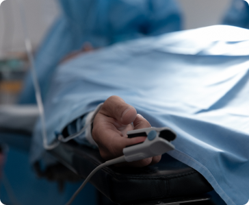 A person lies on a hospital bed covered with a blue sheet, with a pulse oximeter attached to their finger, indicating a medical or surgical setting.