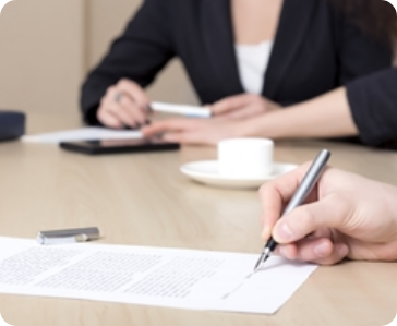 Two people sit at a table during a meeting. One person is writing on a document with a pen, while the other holds a pen and sits near a cup and saucer. Both are dressed in business attire. Two people sit at a table during a meeting. One person is writing on a document with a pen, while the other holds a pen and sits near a cup and saucer. Both are dressed in business attire.