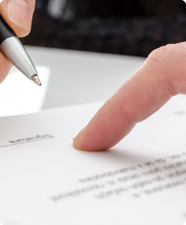 A close-up of a hand holding a pen and a finger pointing to the signature line on a document, preparing to sign. The text on the paper is slightly blurred except for the word "Signature.