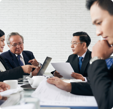 Four business professionals in suits sit at a table with documents, a laptop, and coffee cups, engaged in a serious meeting or discussion in a modern office with a white brick wall background.