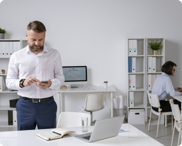 A man in business attire stands at a desk using his phone in a modern office, while a woman works at another desk in the background surrounded by shelves and office supplies.
