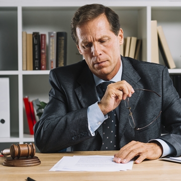 A man in a suit sits at a desk, holding eyeglasses and reviewing documents. A gavel and legal books are on the desk, and shelves with books are in the background, suggesting a legal or office setting. A man in a suit sits at a desk, holding eyeglasses and reviewing documents. A gavel and legal books are on the desk, and shelves with books are in the background, suggesting a legal or office setting.