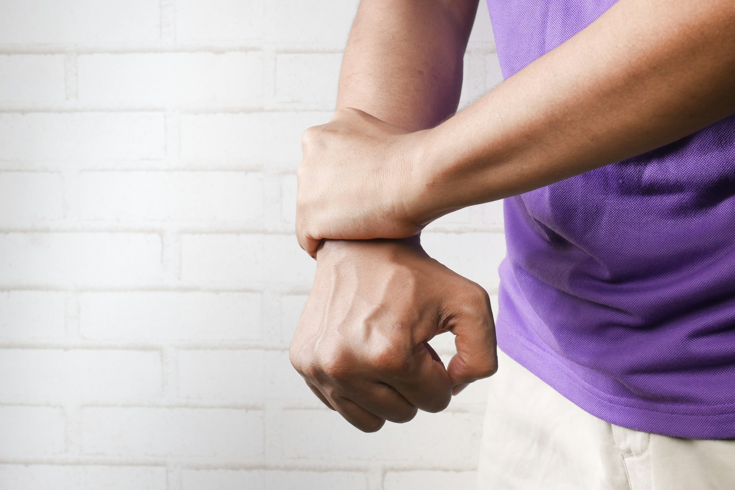 A person wearing a purple shirt and light pants holds their right wrist with their left hand against a white brick wall background. A person wearing a purple shirt and light pants holds their right wrist with their left hand against a white brick wall background.