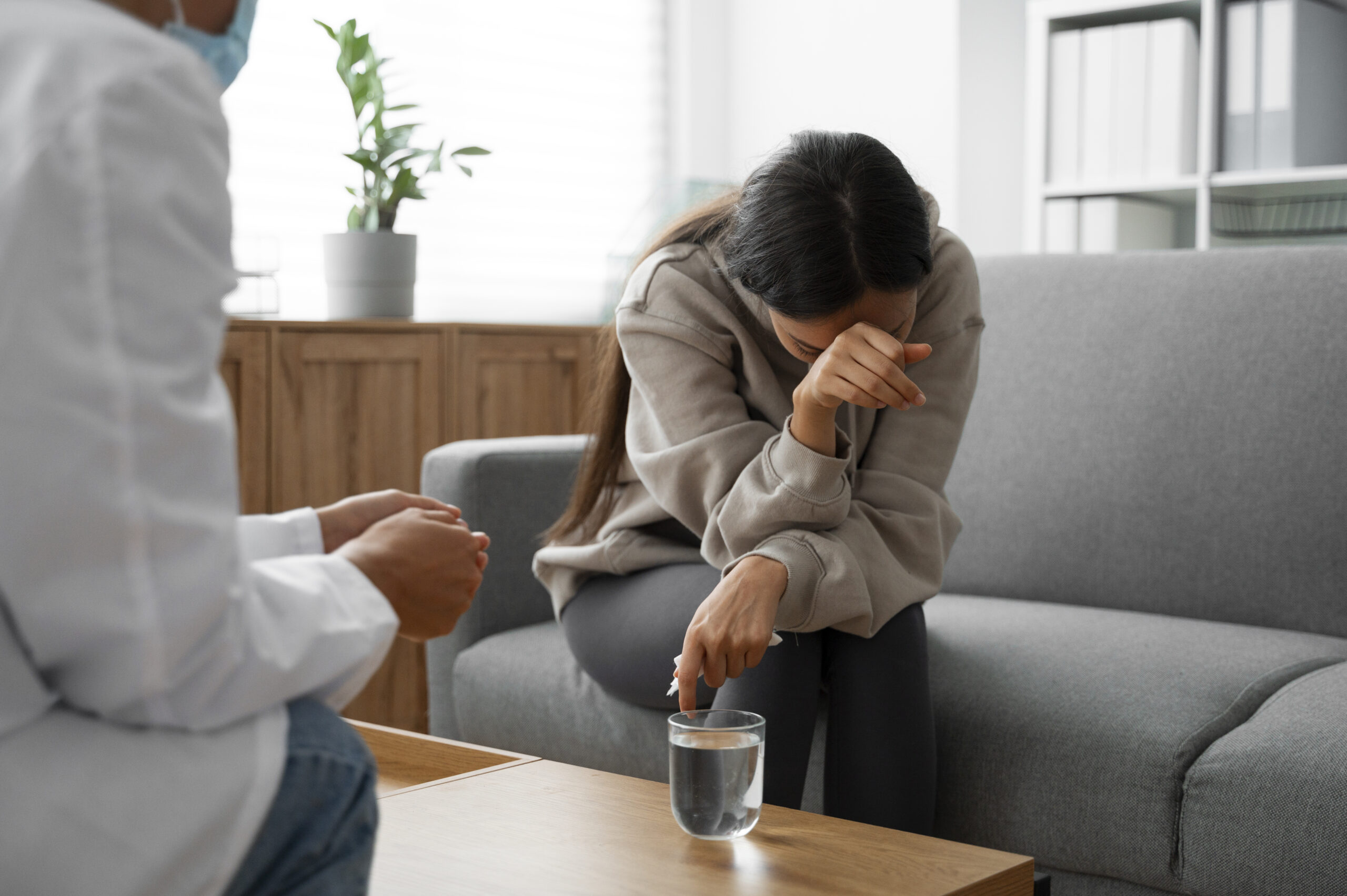 A person sits on a gray couch, visibly upset with their head down and arm covering their face, while another person, likely a professional, sits nearby offering support. A glass of water is on the table in front of them. A person sits on a gray couch, visibly upset with their head down and arm covering their face, while another person, likely a professional, sits nearby offering support. A glass of water is on the table in front of them.
